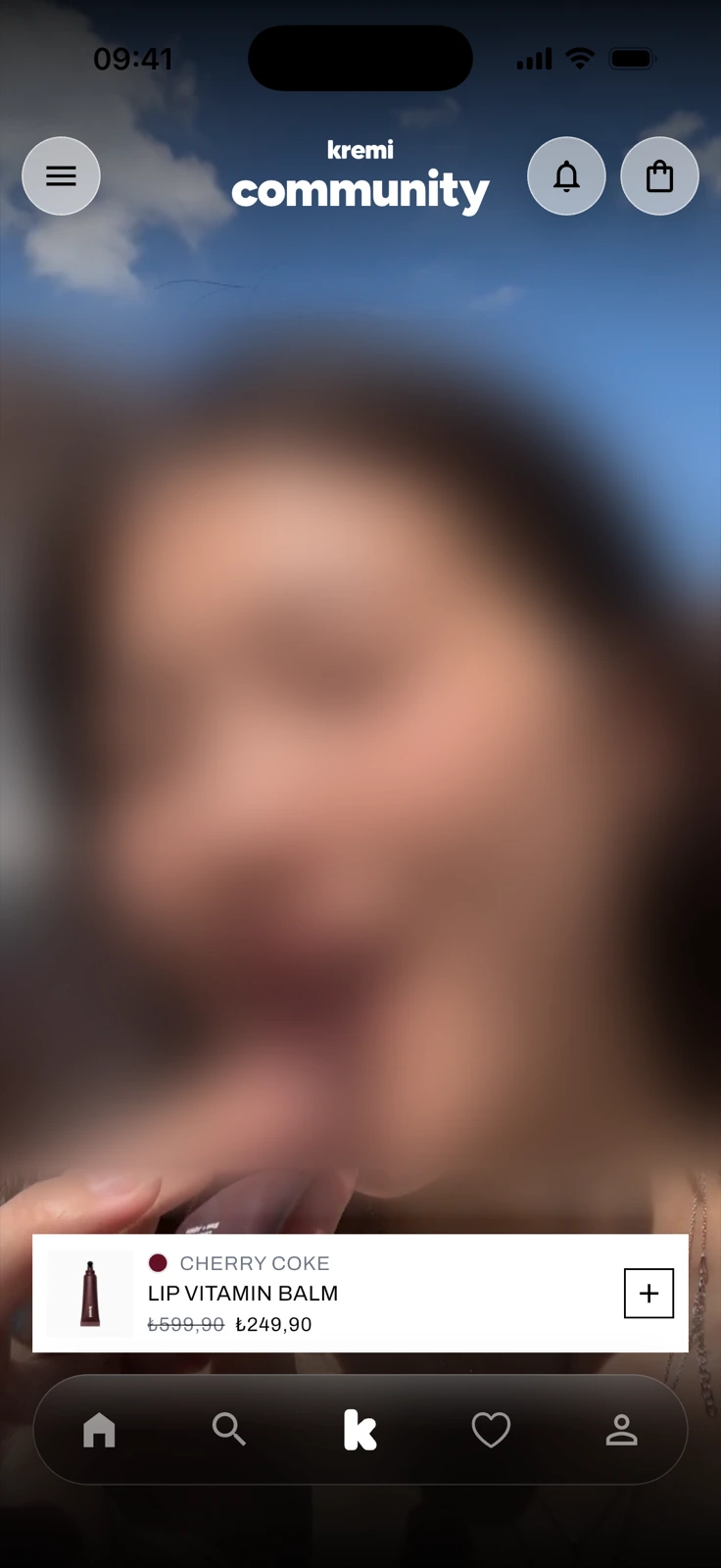The community tab: a full-screen portrait video, deliberately blurred here, under a kremi community header, with a card for Lip Vitamin Balm in the shade Cherry Coke, its old and new price and a plus button, above the tab bar.